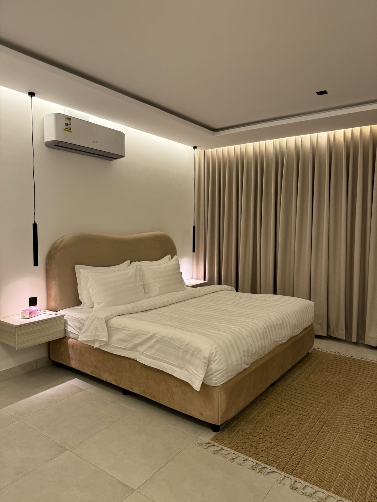 A modern bedroom features a soft, beige bed with crisp white linens. Soft lighting highlights the headboard and the adjacent curtains, which cascade down to the floor. A small bedside table is positioned beside the bed, contributing to the clean, minimalistic design.