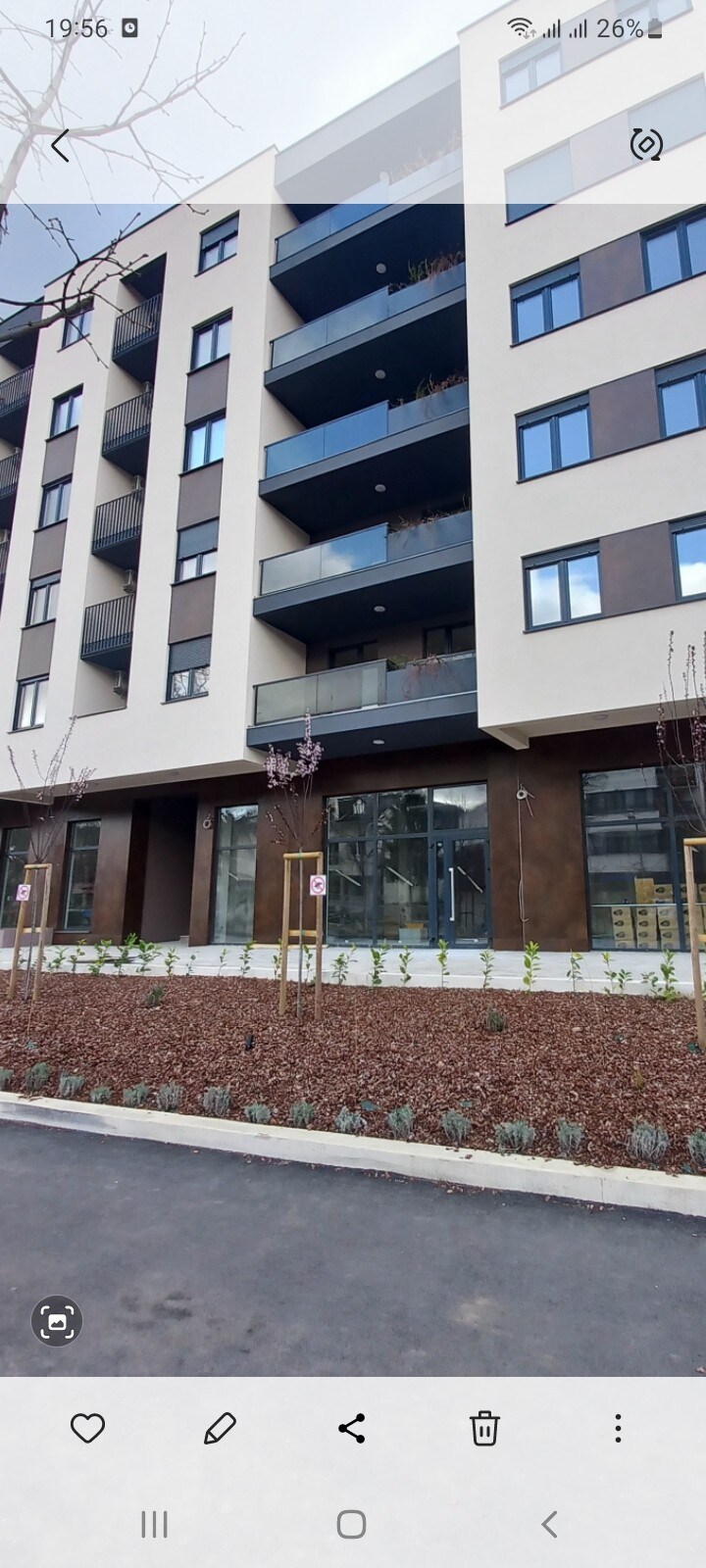A modern building features a clean façade with multiple balconies providing outdoor space. Large windows are present, allowing natural light into the interior. The landscape includes minimalist garden beds with a combination of shrubs and decorative stones, creating a neat appearance in the front area.