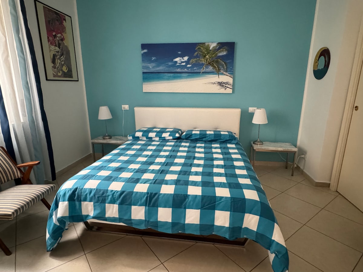 The room features a spacious bed covered with a checkered blue and white blanket. A painting depicting a beach scene is hung above the bed. Two bedside tables with lamps are positioned on either side, and a comfortable chair is visible in the corner.