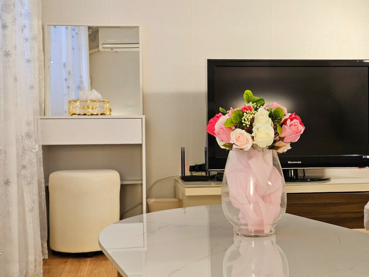 A modern living area is presented, featuring a glass vase filled with an arrangement of artificial flowers in soft pink and white tones. A television sits on a media unit, with a sleek mirror and a small ottoman visible in the background.
