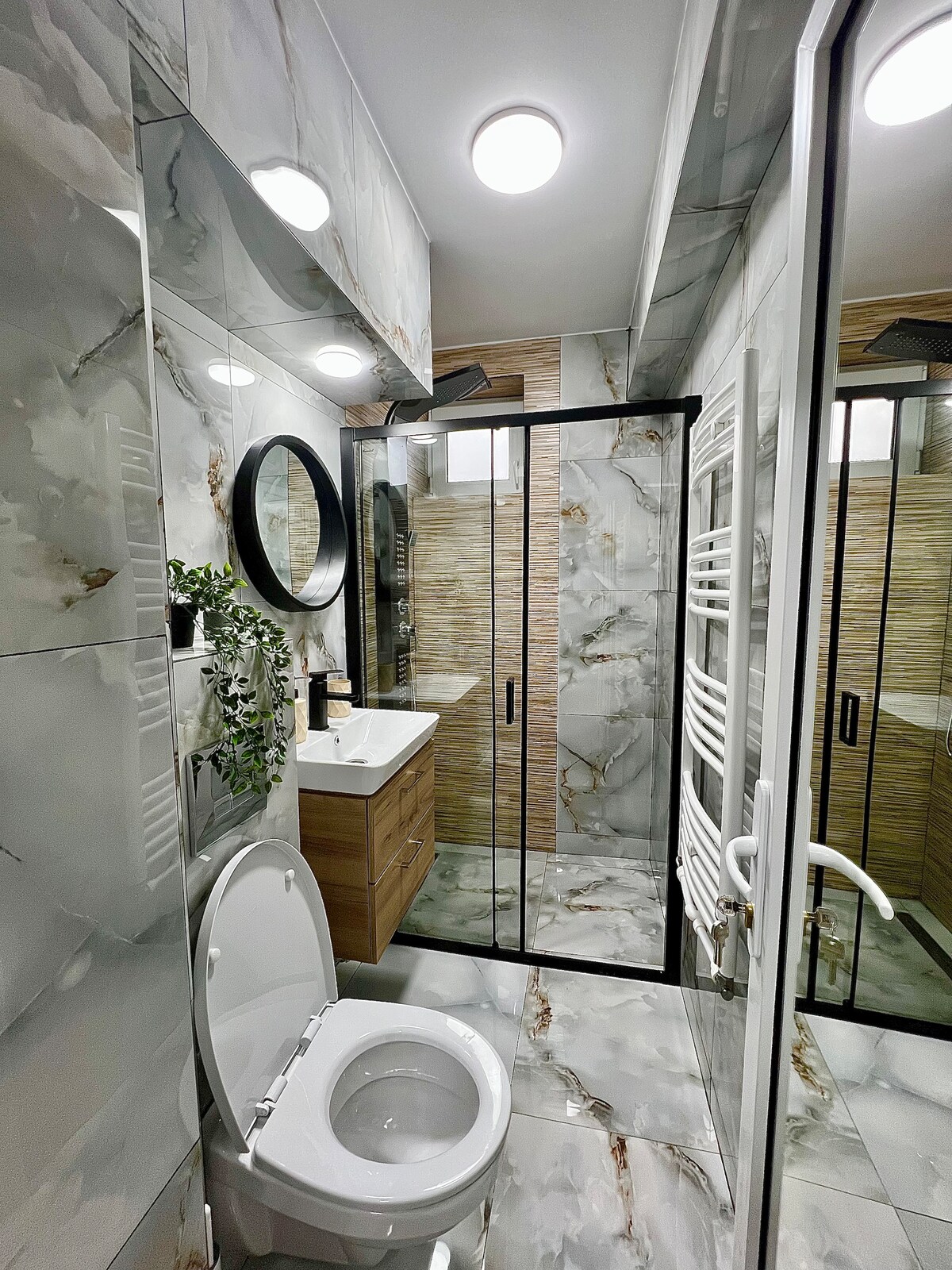 A modern bathroom is shown, featuring stylish marble tiles and a spacious walk-in shower with glass doors. A sleek vanity with a circular mirror is positioned above a wooden cabinet. Bright overhead lights enhance the contemporary design, while a towel warmer is visible on the wall.