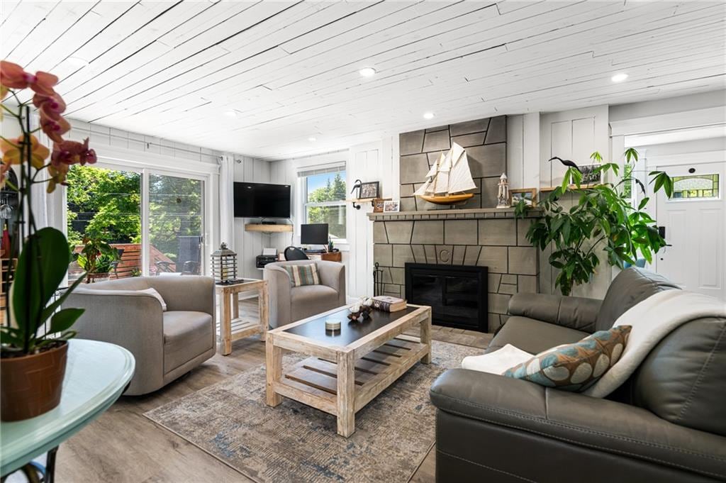 A cozy living room is showcased with a stone fireplace as the focal point. Comfortable seating arrangements include a leather sofa and armchairs. A coffee table with a rustic finish sits in the center, while natural light streams in through large windows adorned with light curtains.