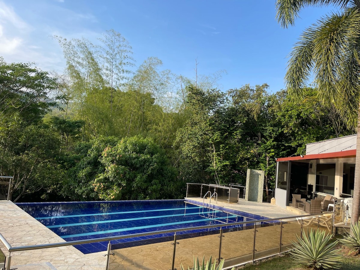 An outdoor pool is surrounded by lush greenery, with blue tiles reflecting the sky. A lounge area is visible nearby under a covered patio, featuring comfortable seating and a dining table. The relaxing space is enhanced by the natural landscape.