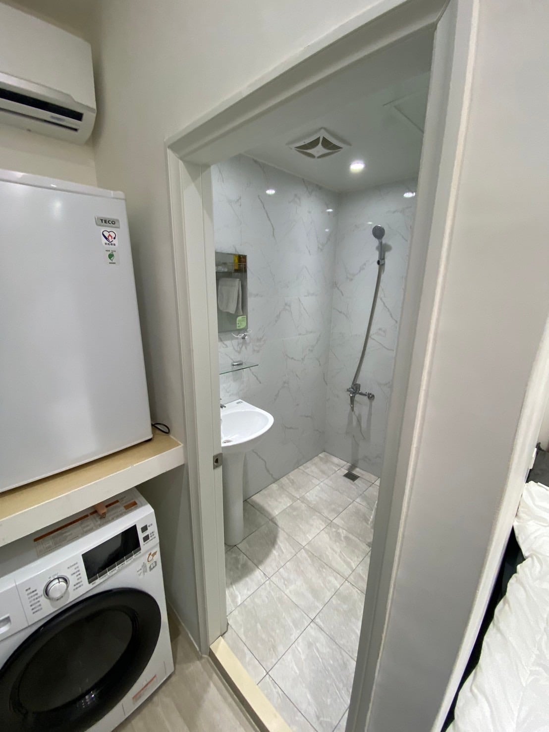 A compact bathroom area is visible, featuring a standing shower with a glass partition and a modern sink. The walls are adorned with white marble accents, and ambient lighting is provided by recessed ceiling lights. A washing machine is located beside the bathroom door.