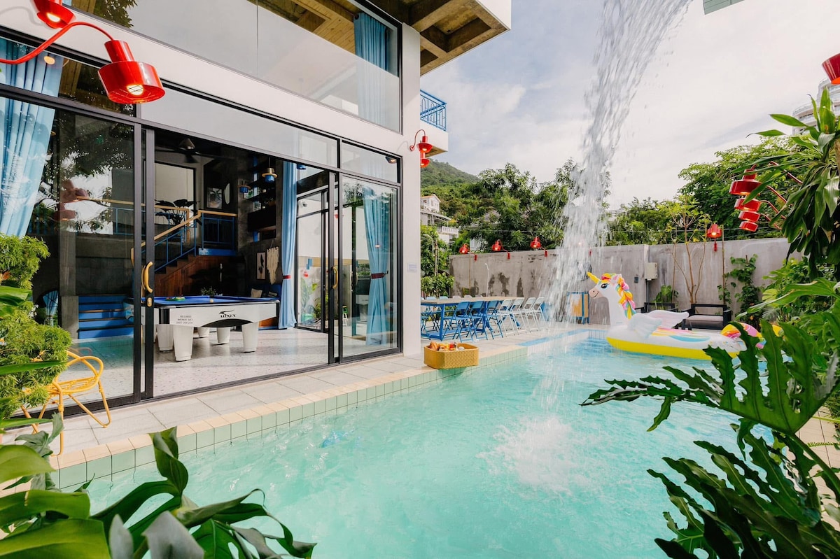 A spacious outdoor area features a 40 square meter swimming pool with a led-lit waterfall, surrounded by lush greenery. Large glass doors open to a sunlit lounge, while colorful seating and a floating inflatable add to the inviting space.