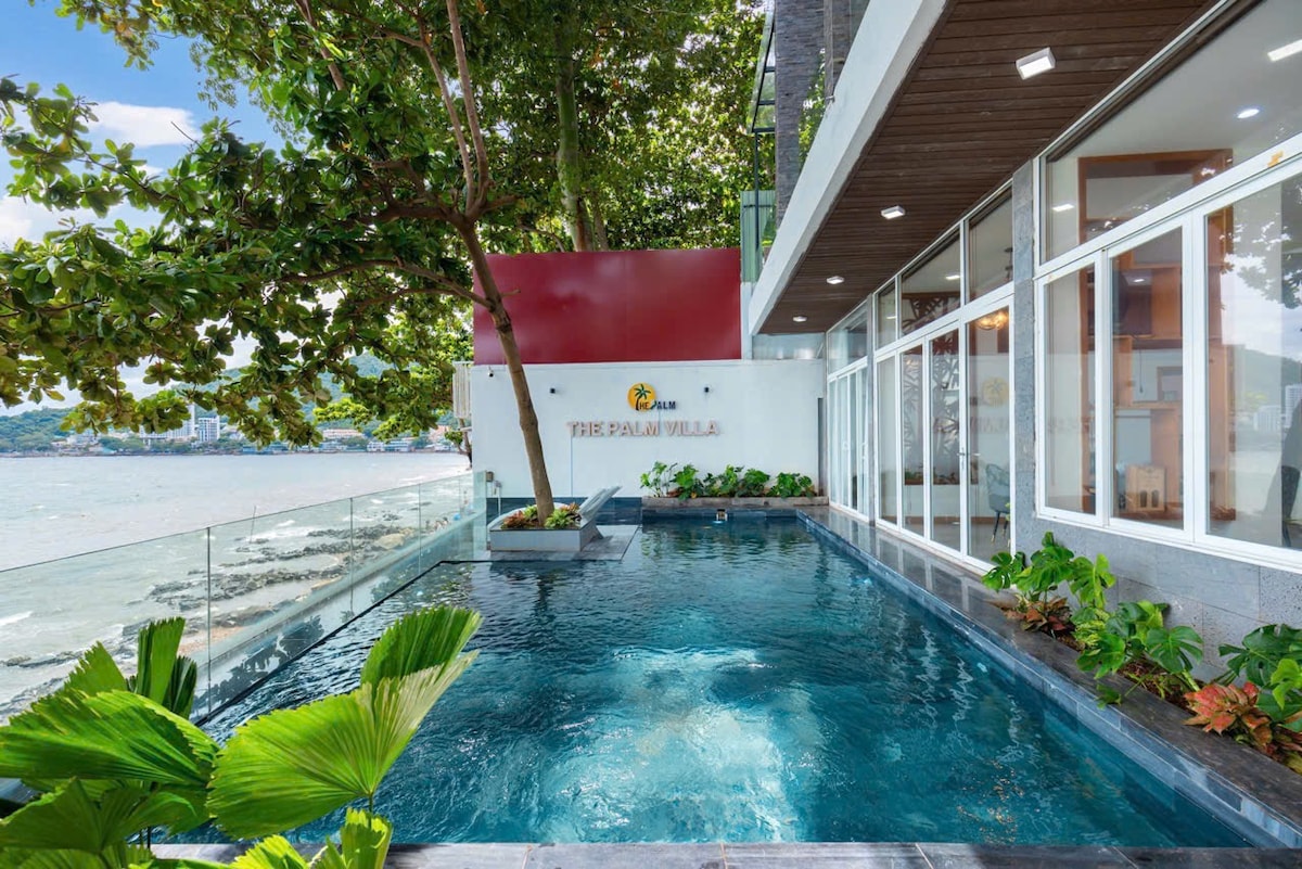 A refreshing pool is situated beside a modern building, framed by greenery and a clear view of the ocean. Sunlight reflects off the water's surface, creating a serene ambiance. Large windows offer glimpses into the interior, enhancing the connection between indoor and outdoor spaces.