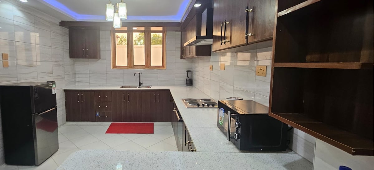 The kitchen features modern cabinetry with a stone countertop, complemented by a sleek refrigerator and a microwave. A four-burner stove is positioned next to the sink, with a window providing natural light. A red mat adds a touch of color to the tiled floor.