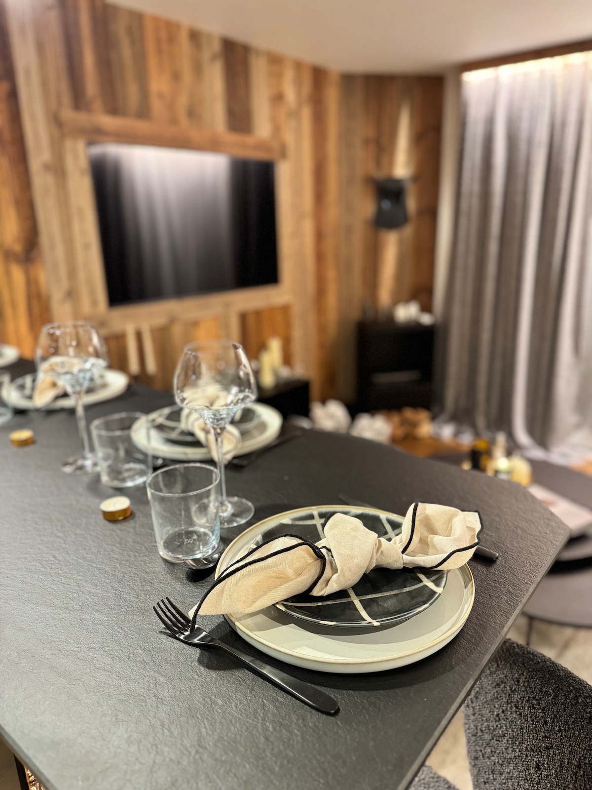 A dining setup is presented with elegant tableware, featuring plates, wine glasses, and cutlery. The backdrop includes wooden paneling and a TV, with soft lighting from a nearby window. Textured curtains lend a cozy element to the atmosphere.
