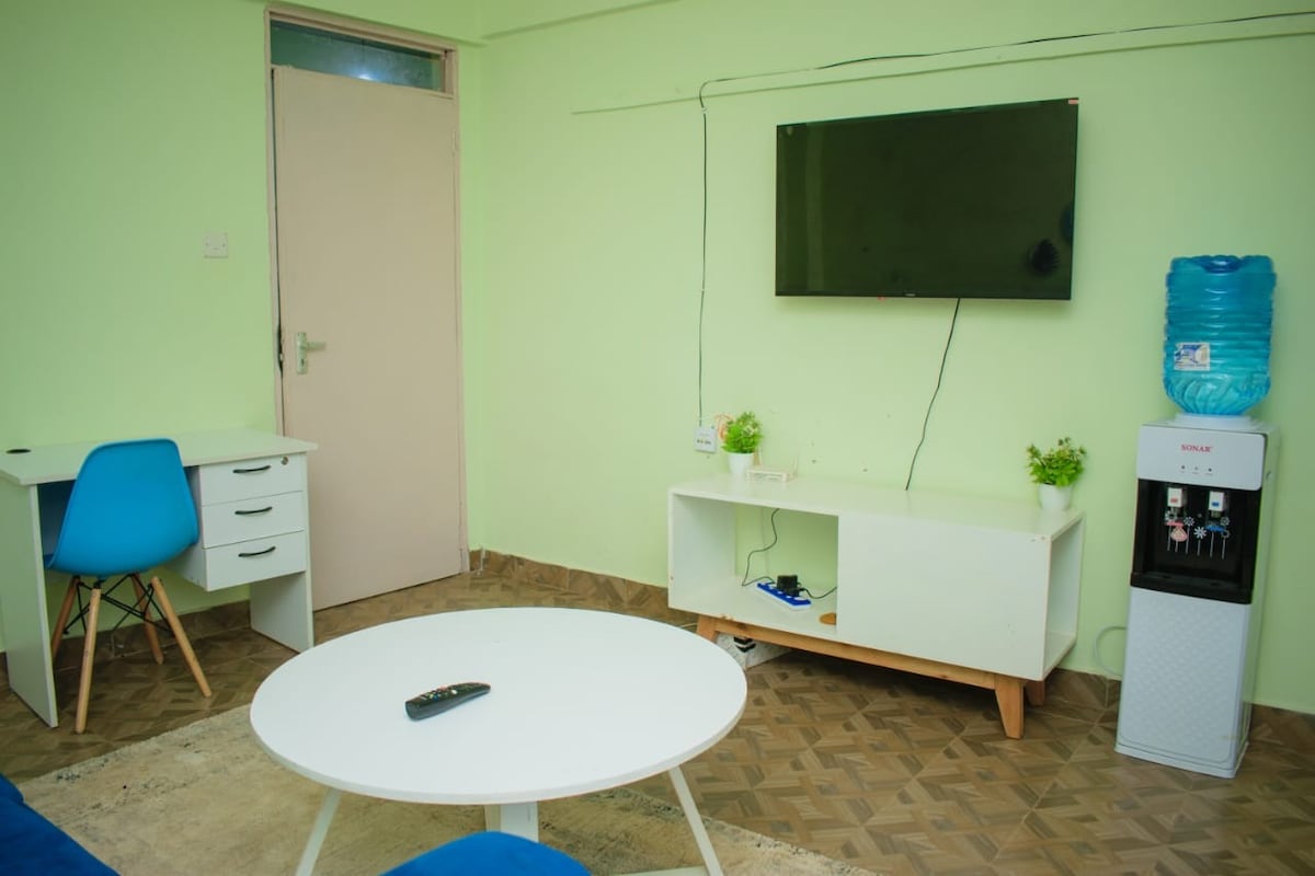 A bright and airy living area features light green walls, a white coffee table, and a desk with a blue chair. A television is mounted on the wall, and a water cooler is positioned nearby, adding to the functionality of the space.