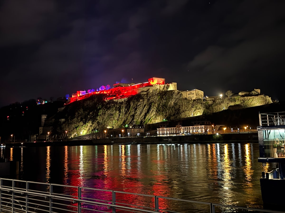 A lit-up fortress is positioned on a hillside, creating a striking contrast against the dark night sky. Its warm lights reflect off the water's surface below. Along the riverbank, structures and walkways can be seen, adding to the serene riverside atmosphere.