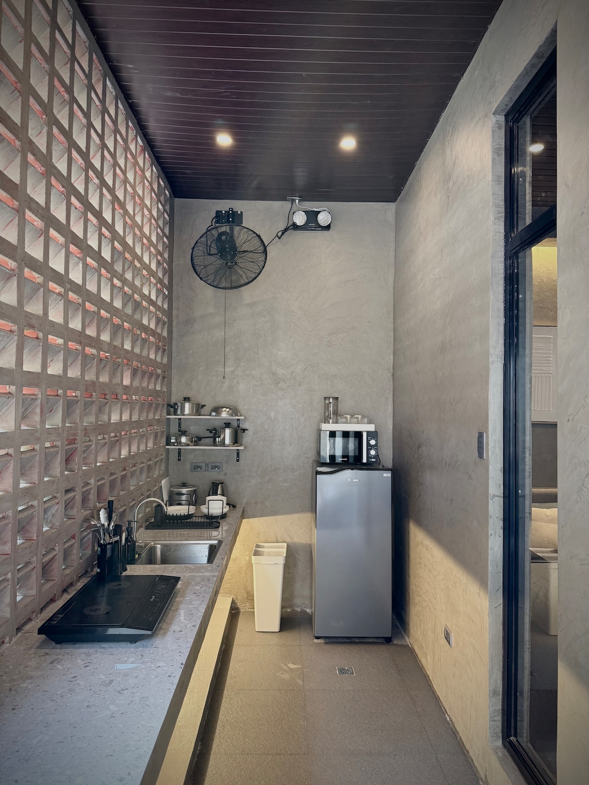 A minimalist outdoor kitchen features a sleek countertop and a modern stove. A blended gray aesthetic includes a compact refrigerator and wall-mounted shelves for dinnerware. Natural light pours in from a large window, enhancing the functional yet simple design.