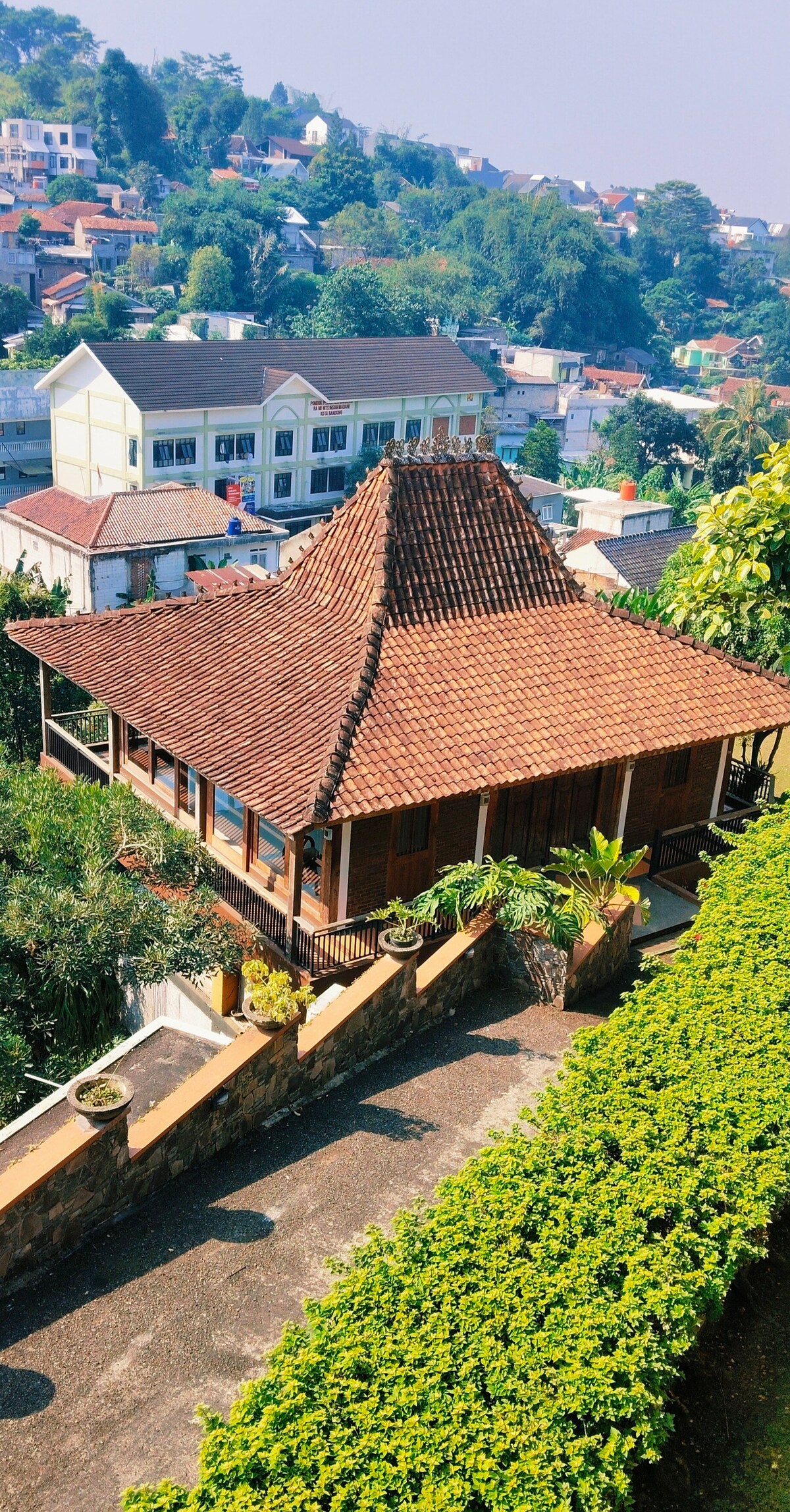 An intricately designed joglo house is showcased from an elevated viewpoint. The traditional roof features distinct triangular peaks, surrounded by lush greenery. The house rests on a stone foundation, framed by neatly trimmed hedges and various potted plants that enhance the inviting outdoor space.