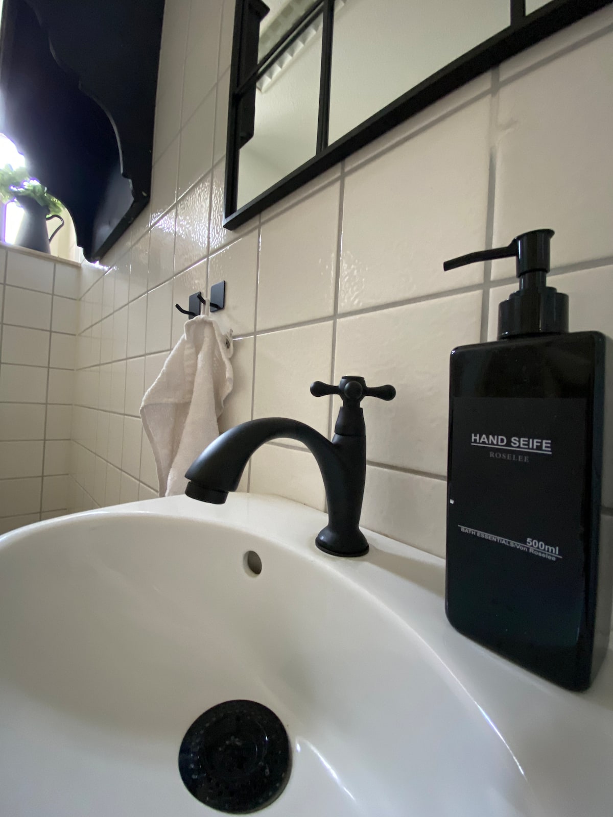 A modern bathroom sink is highlighted, featuring a sleek black faucet and a dark soap dispenser. A white towel hangs on a nearby hook, with a mirror reflecting the arrangement. Subtle natural light filters in from a window above, enhancing the clean lines of the design.