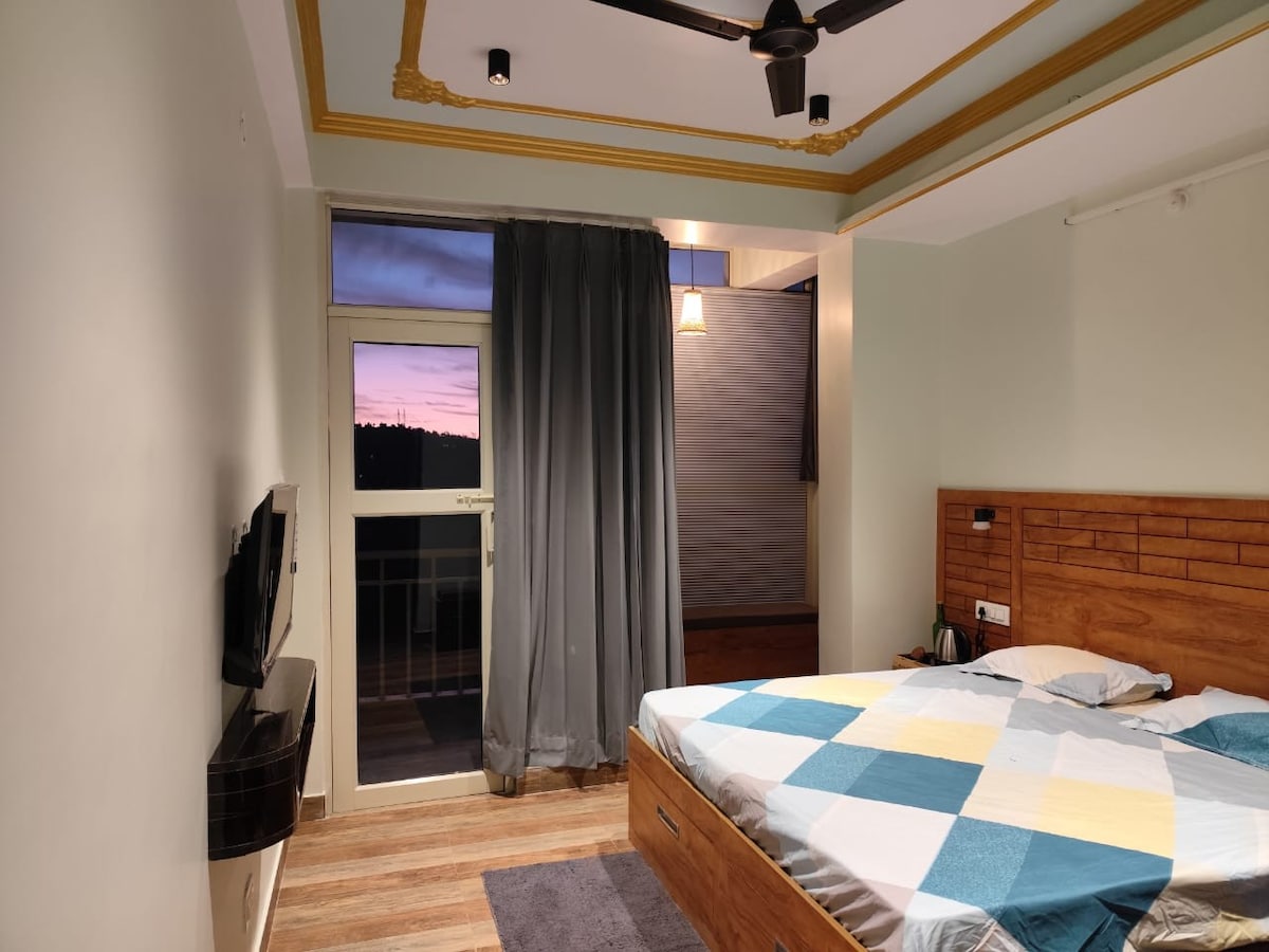 A well-lit bedroom features a spacious bed with a patterned blanket. A mounted television is visible on the wall opposite the bed. Sheer curtains frame a balcony door that opens to a sunset view, while decorative ceiling elements add character.
