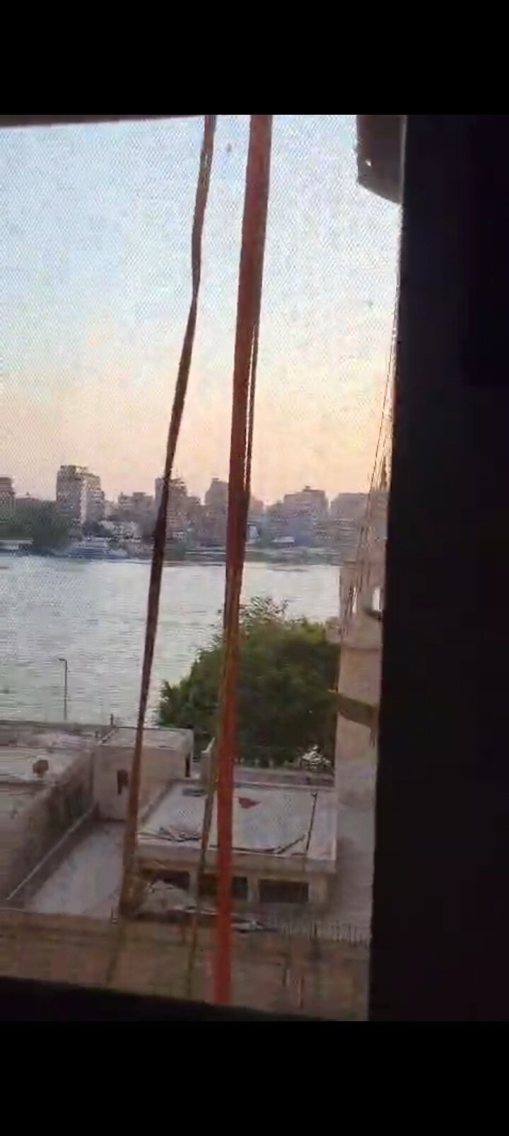 A view of the river and city skyline is captured through a window screen. The soft hues of sunset reflect on the water, while nearby structures are visible. Thin, colorful ribbons hang from the window frame, adding a touch of local character.