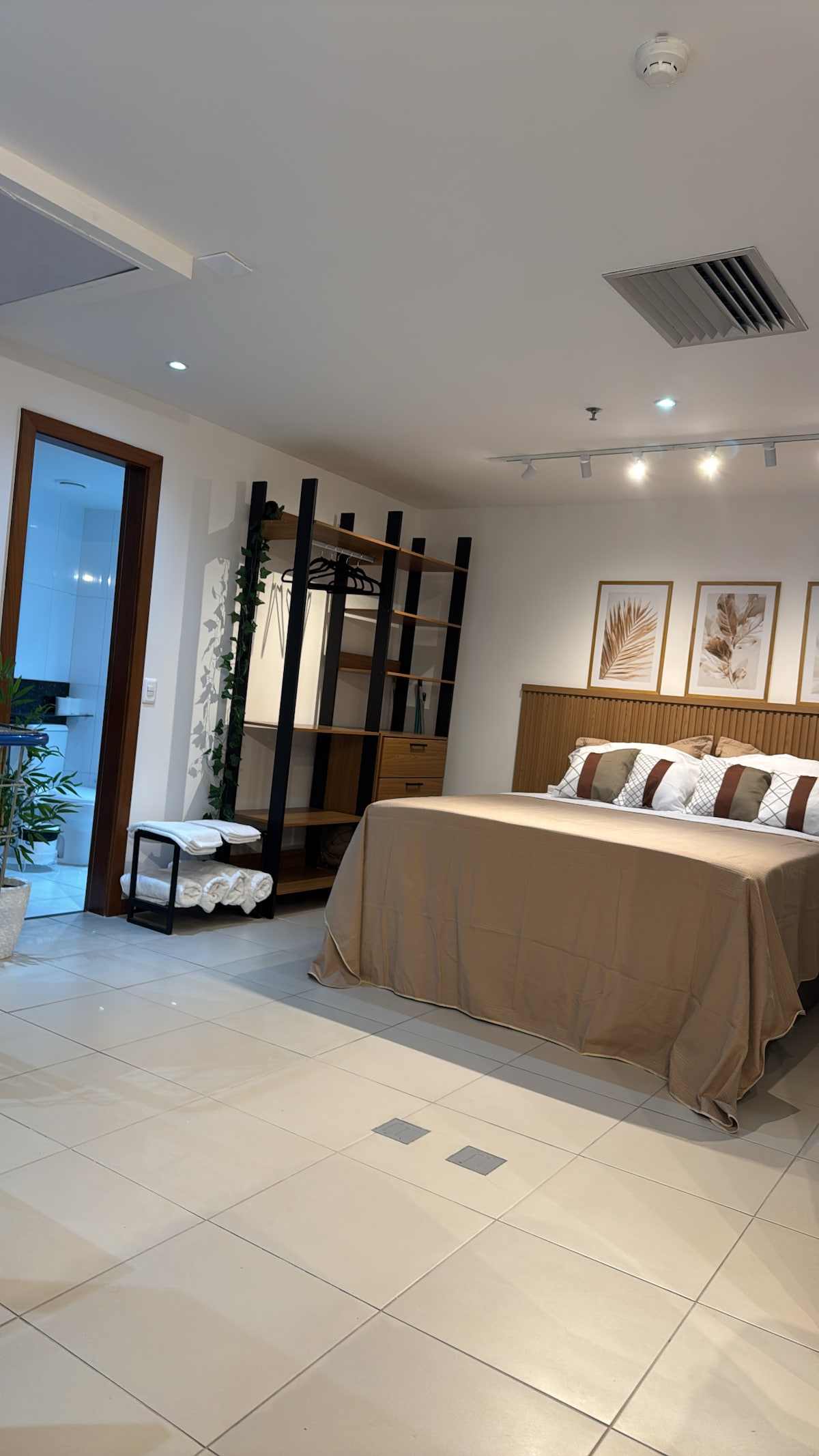 A contemporary bedroom features a spacious bed with neutral-colored linens, complemented by decorative pillows. A tall shelving unit stands against the wall, accompanied by a small storage cart. Natural light enters through the doorway leading to a bathroom, enhancing the serene atmosphere.