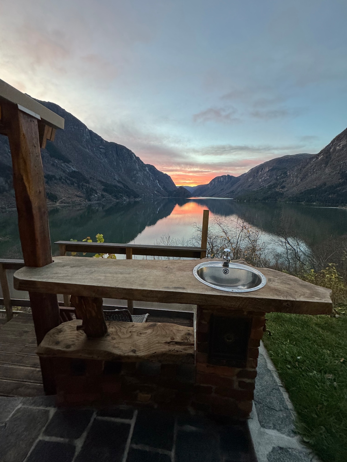 A wooden countertop with a sink is positioned outdoors, offering a view of the scenic lake and surrounding mountains. The sunset casts warm colors across the sky, reflecting on the water. The area is framed by gently sloping hills, creating a serene atmosphere.