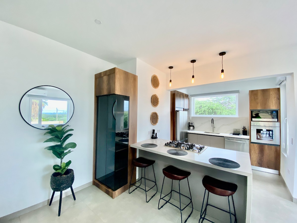 A modern kitchen features sleek cabinetry and a central island with bar seating. Pendant lights hang above, illuminating the countertops. Large windows provide natural light and showcase the lush outdoor views. A green plant in a decorative pot adds a touch of nature to the space.