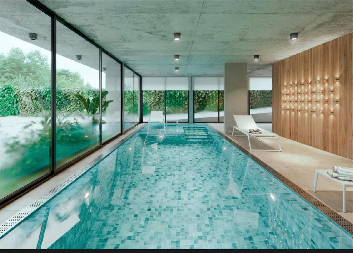 An indoor swimming pool is surrounded by expansive glass walls, allowing natural light to fill the space. A serene atmosphere is created by the smooth stone tiles and modern wooden accents. Sun loungers are positioned neatly beside the pool, enhancing the relaxation area.