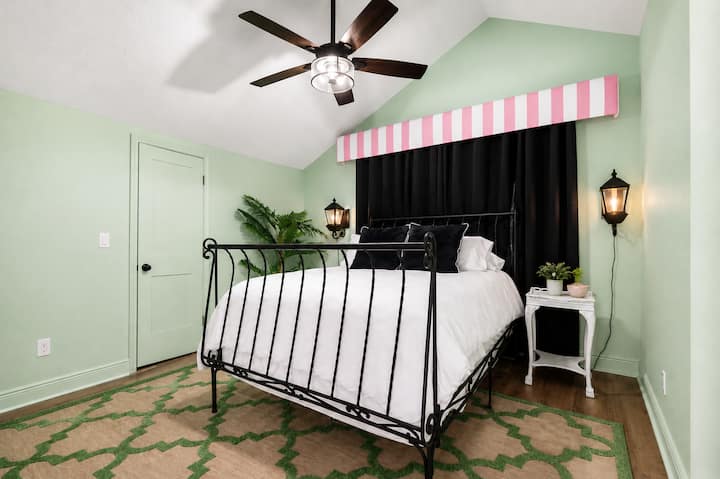 Relax in this peaceful queen
 bed featuring soft mint-green walls, a black iron bed, blackout curtains, vaulted ceilings, cozy lighting, and tropical-inspired accents. A stylish retreat designed for a restful night's sleep after a fun day in St. Aug