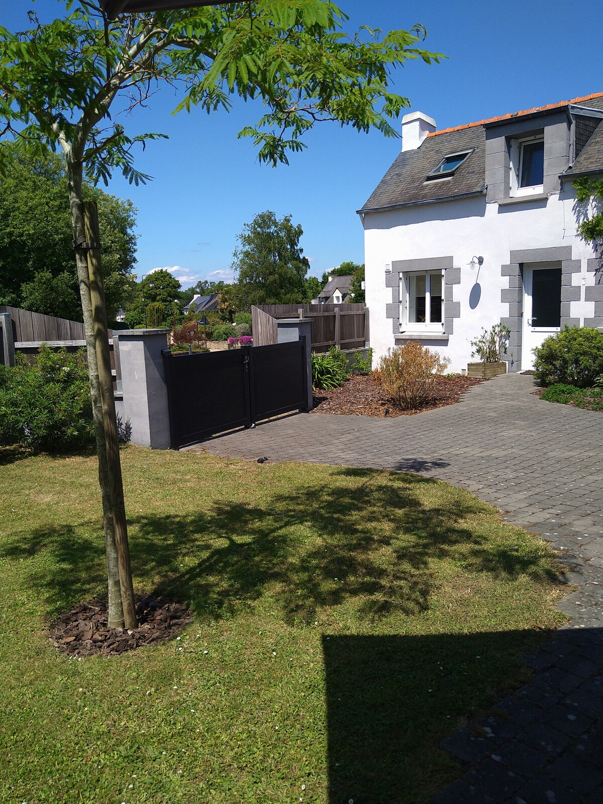 The exterior of the house is presented, featuring a manicured garden with a young tree and flowerbeds alongside the pathway. A black gate provides access to the property, while the charming façade of the house is visible under a clear blue sky.