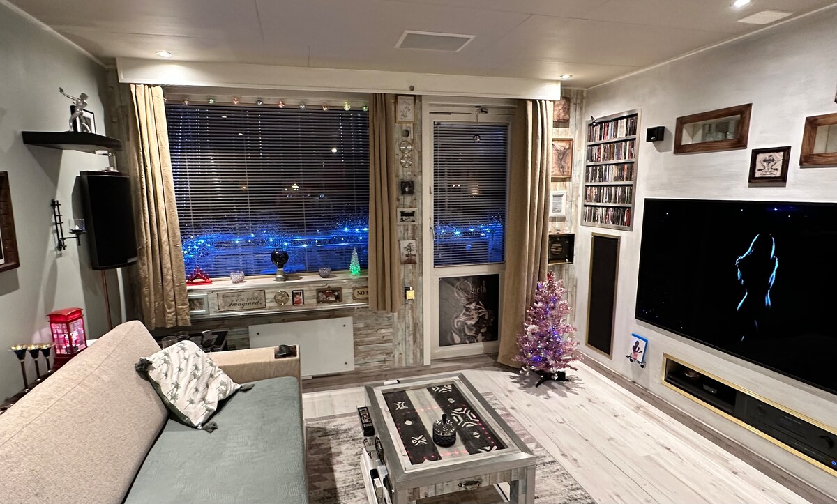 A cozy living area features a light-colored sofa and a coffee table, with shelves displaying various decorative items. A small Christmas tree adds a festive touch. The large window reveals a city view adorned with soft blue lights at night, while a flat-screen TV is mounted on the wall.