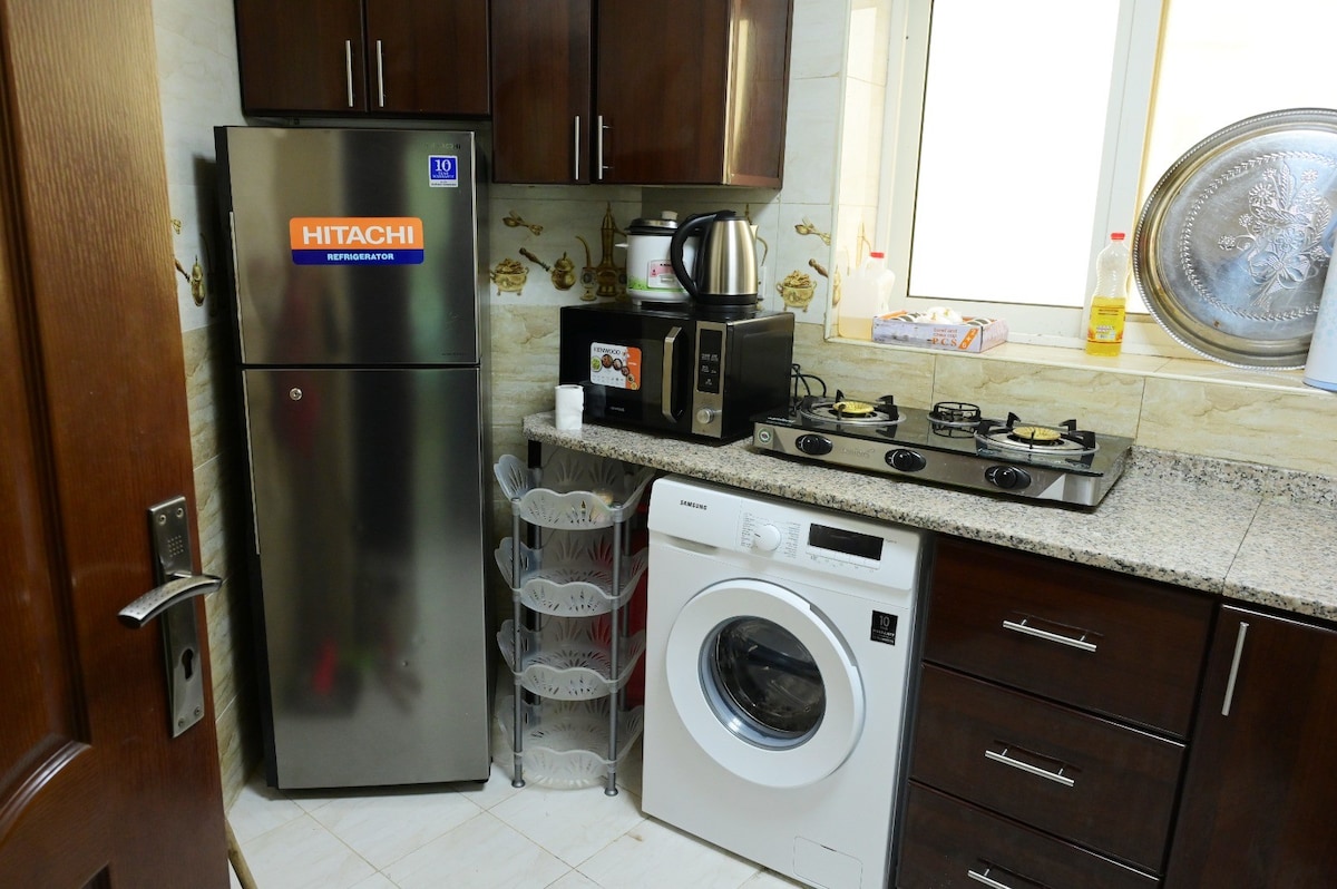 A functional kitchen is presented, featuring a stainless steel refrigerator and a washing machine. A countertop with a coffee maker and a four-burner gas stove is visible, along with a storage rack. Natural light enters through a nearby window.