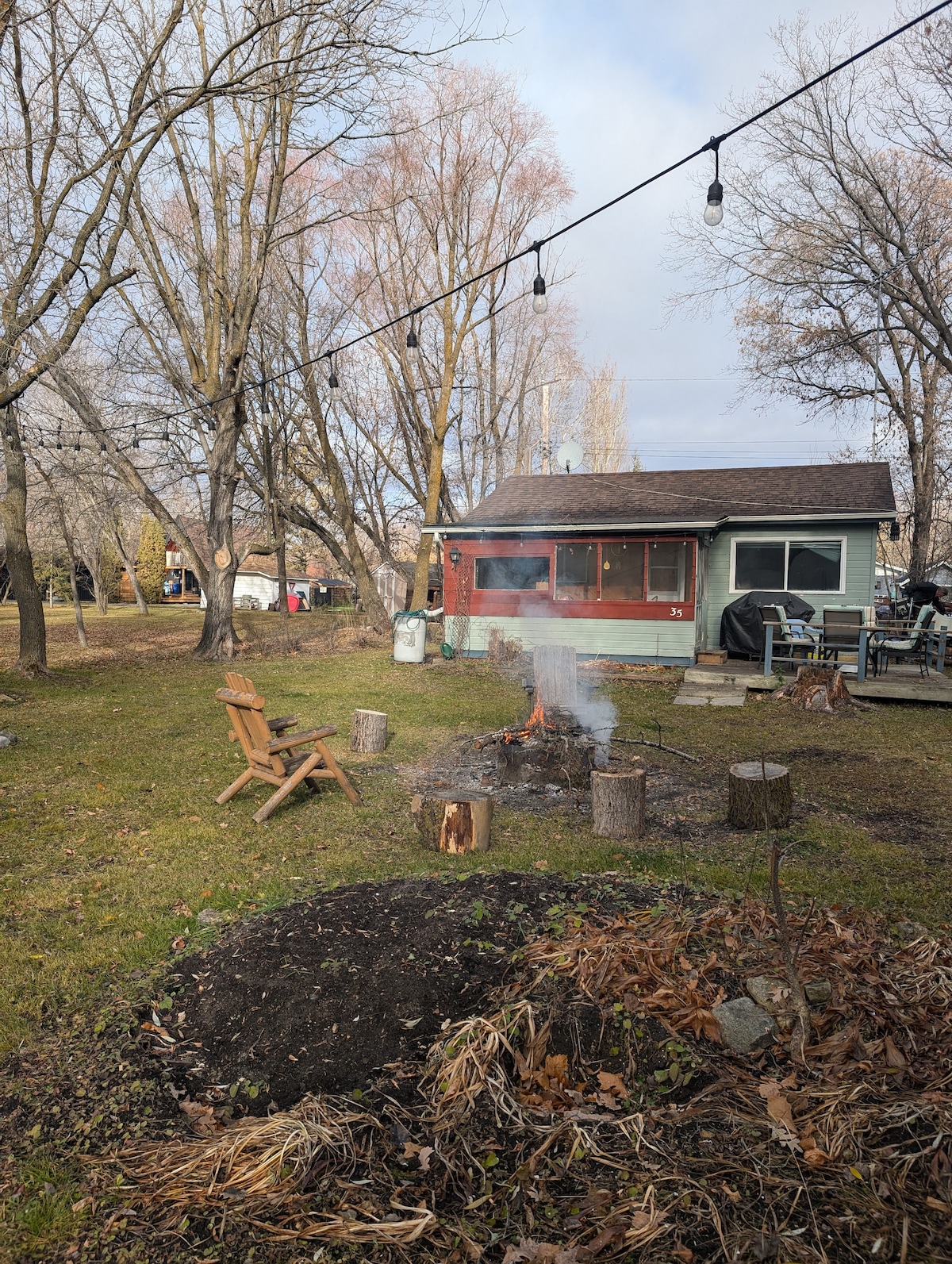 An outdoor seating area features an Adirondack chair next to a fire pit surrounded by logs. A cozy cabin is visible in the background, with trees lining the property. Soft strings of lights hang above, illuminating the yard.