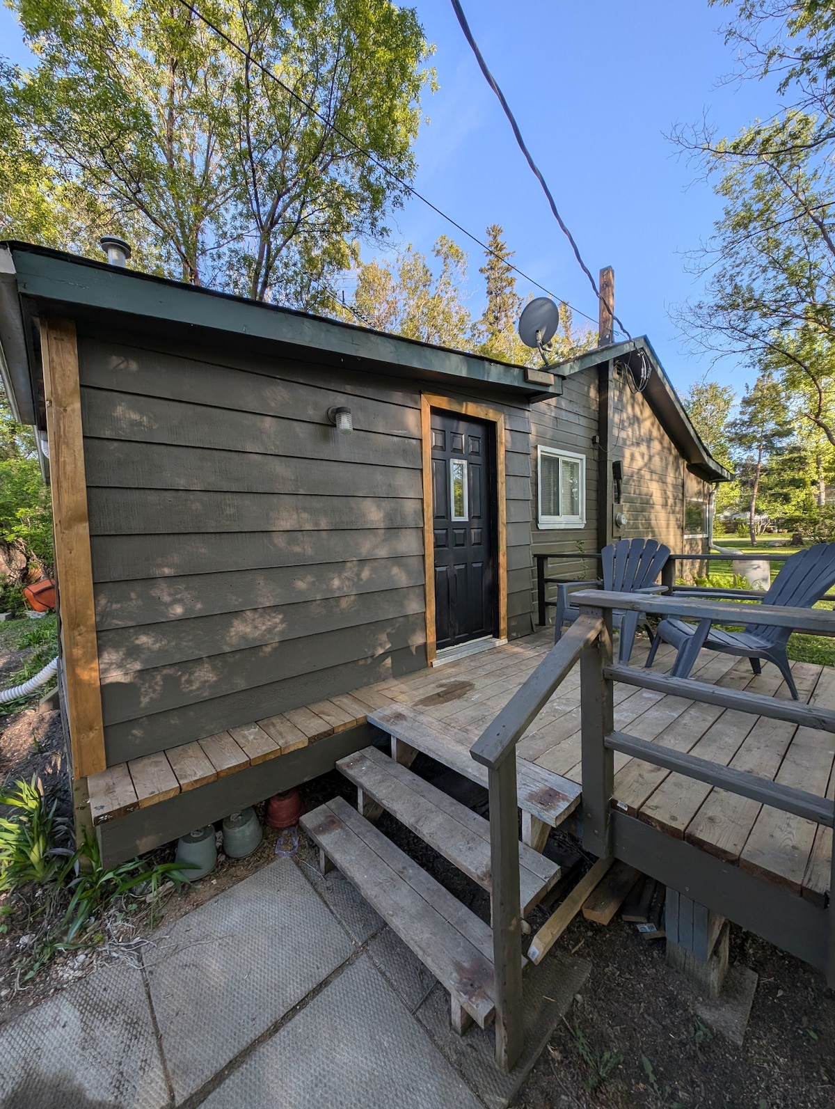 An exterior view of the cabin is presented, featuring a wooden porch with steps leading to a dark front door. Two adirondack chairs are positioned on the deck, surrounded by greenery. Sunlight filters through the trees, casting gentle shadows on the cabin's exterior.