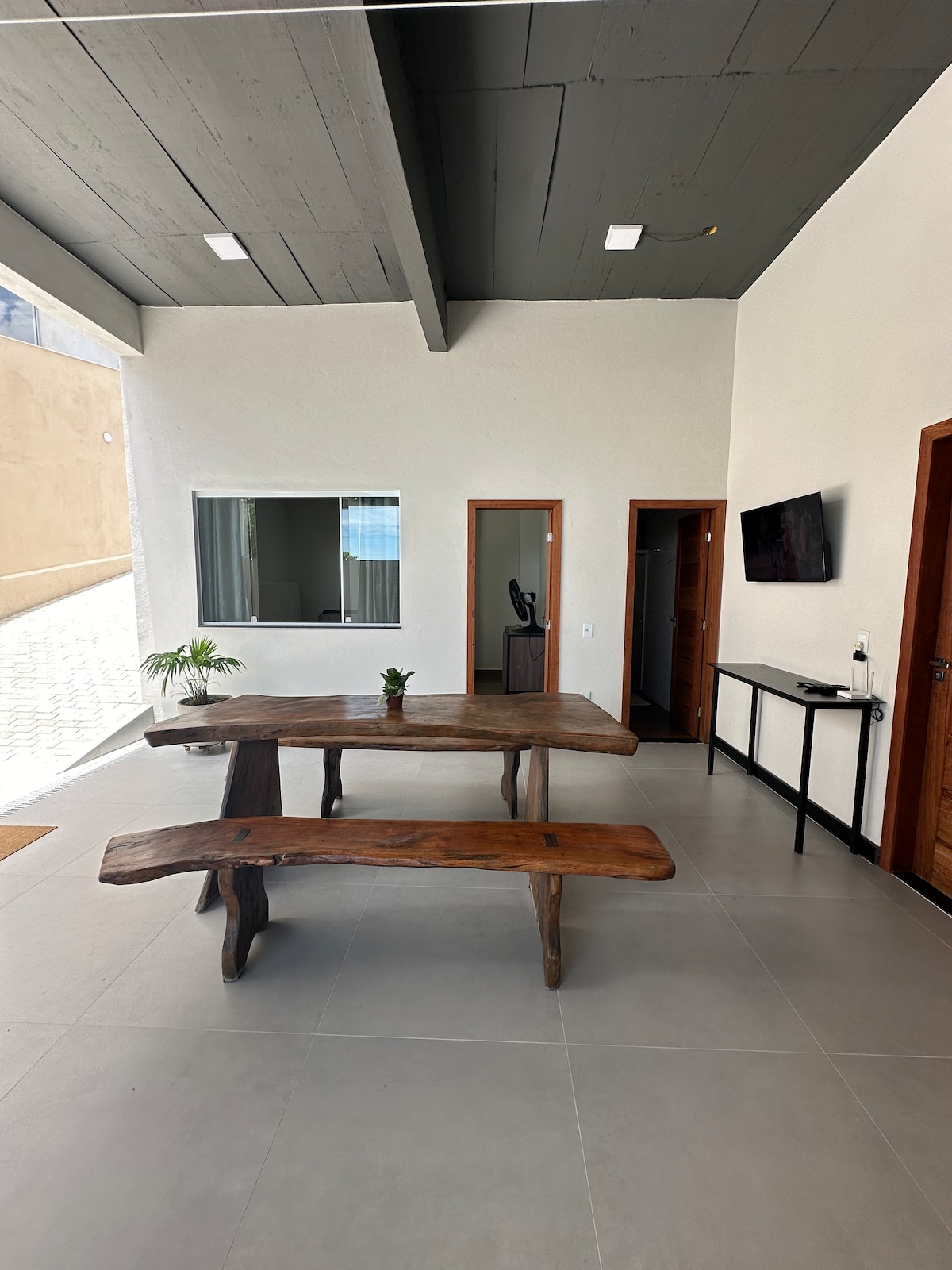 A spacious entryway is displayed, featuring a rustic wooden table with matching benches. Adjacent to the table, a television is mounted on the wall. Natural light is allowed in through large windows. The floor is finished with light tiles, enhancing the airy atmosphere.