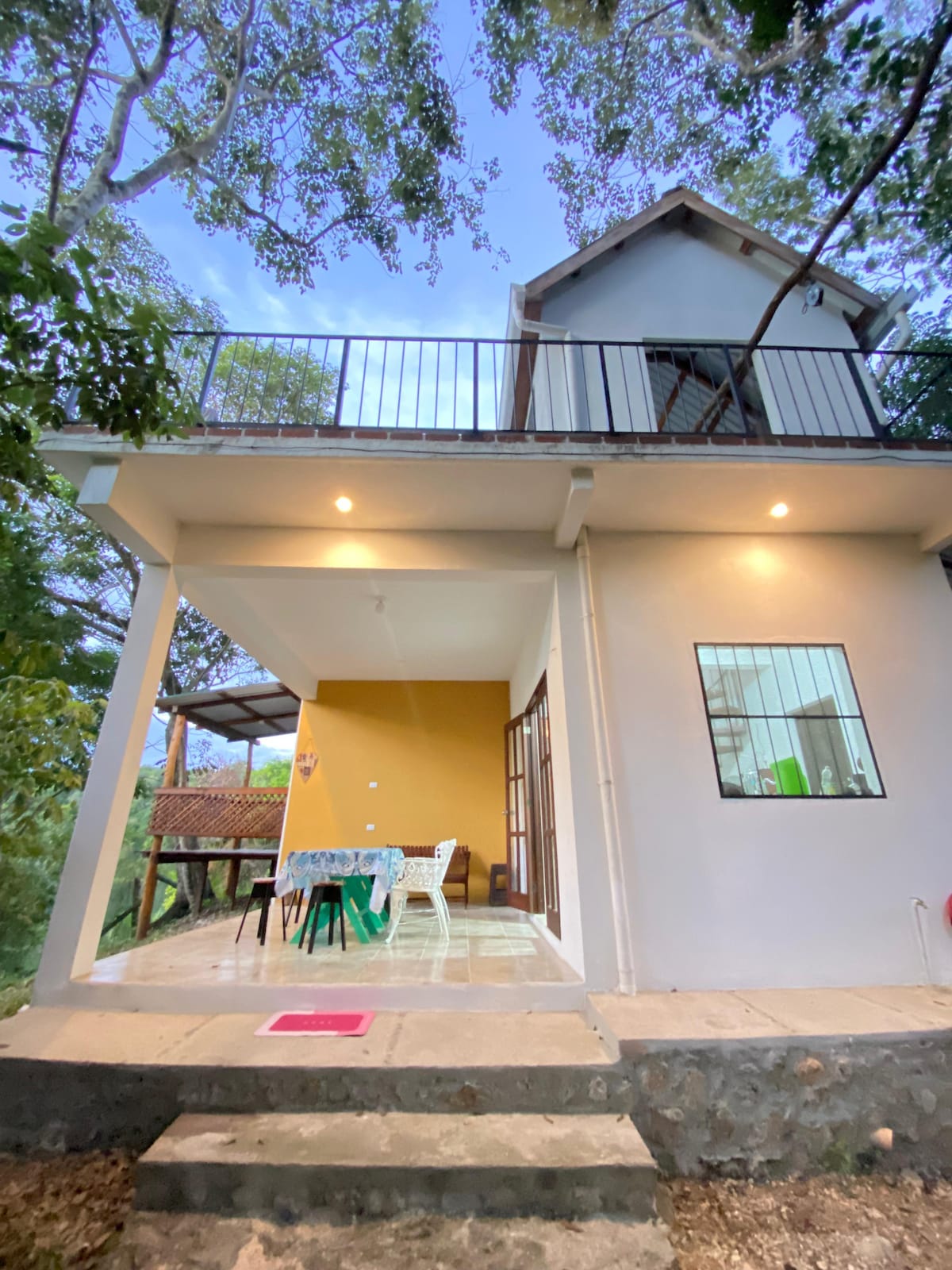 A two-story house is set amidst lush greenery, featuring a spacious terrace with outdoor seating. The exterior combines light-colored walls with a vibrant yellow accent, while large windows provide ample natural light. Steps lead up to the entrance, enhancing accessibility.