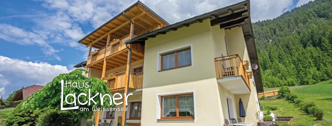 A charming multi-level building is depicted, featuring a light yellow exterior complemented by a wooden balcony on the upper level. Lush greenery surrounds the property, enhancing its serene environment, while a clear sky adds to the welcoming atmosphere.