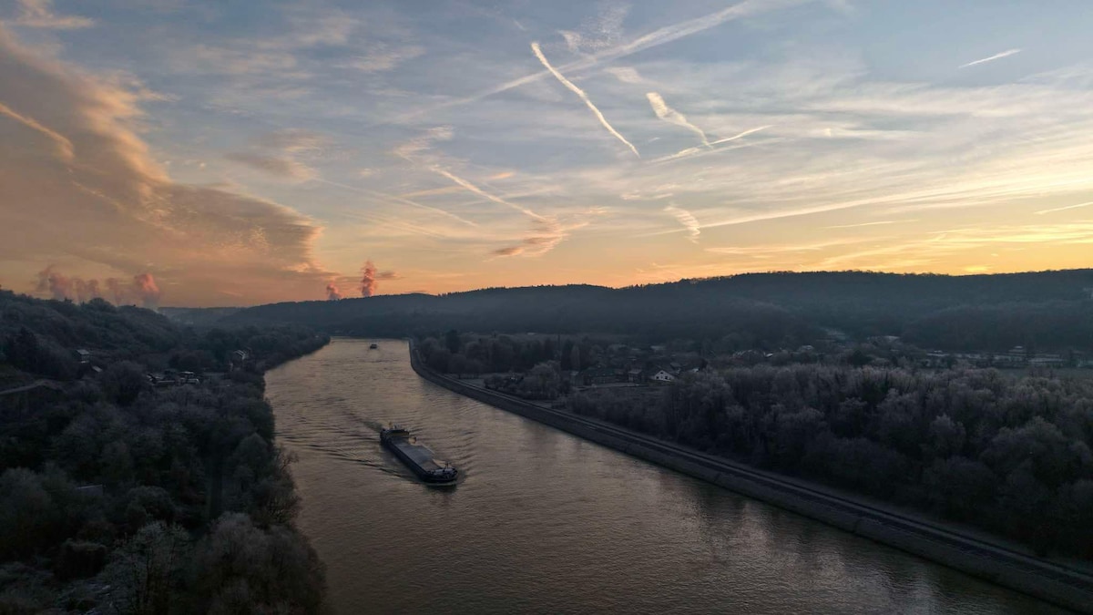 A serene view of the Meuse River is captured at dawn, featuring mist rising over the water. A barge moves gracefully along the river, flanked by tree-covered hills, while soft pastel colors fill the sky, suggesting the start of a peaceful day.