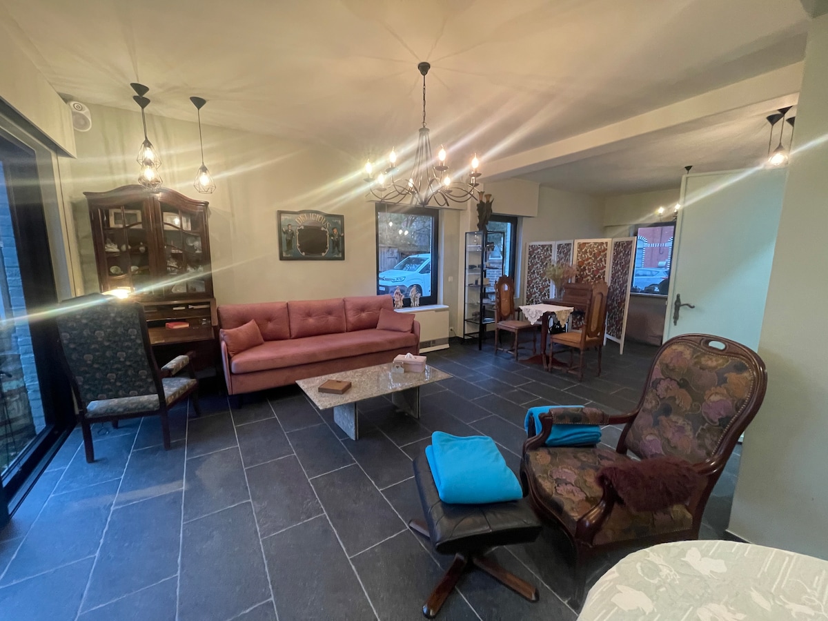 A spacious living area features a pink sofa paired with an intricately designed armchair. A slate floor is complemented by a glass coffee table at the center. Dining chairs surround a small table in the background, with framed artworks adorning the walls.