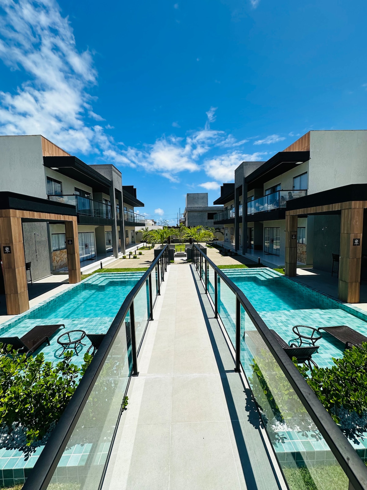 A central pathway links modern two-story buildings, flanked by serene pools. Lush greenery and lounge chairs are visible along the edges, enhancing accessibility. Bright blue skies and soft clouds create a vibrant backdrop above the well-maintained grounds.