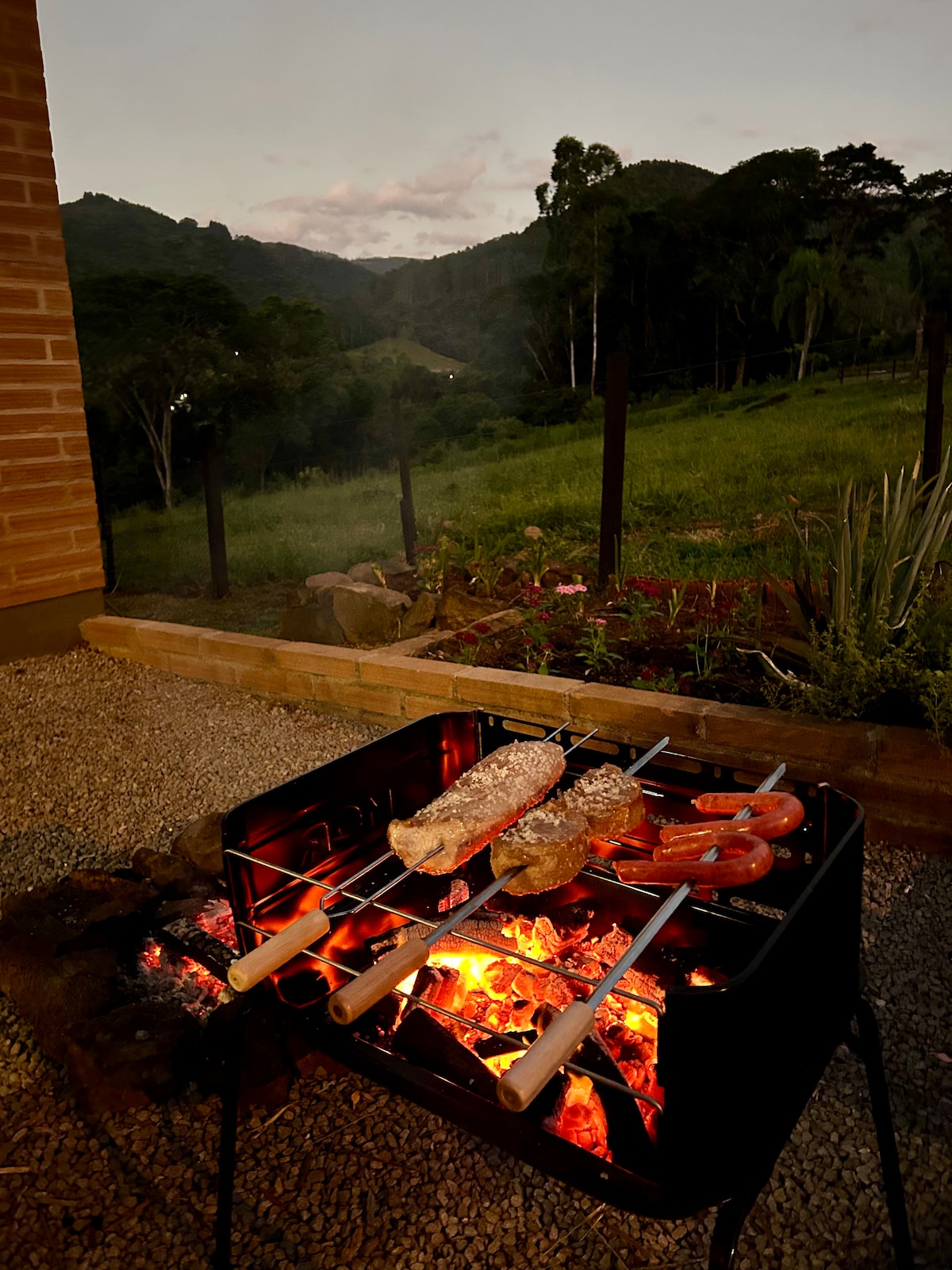A portable grill is set up outdoors, featuring skewers with meat and vegetables placed over glowing embers. In the background, rolling green hills are visible under a twilight sky, creating a serene setting for outdoor dining.