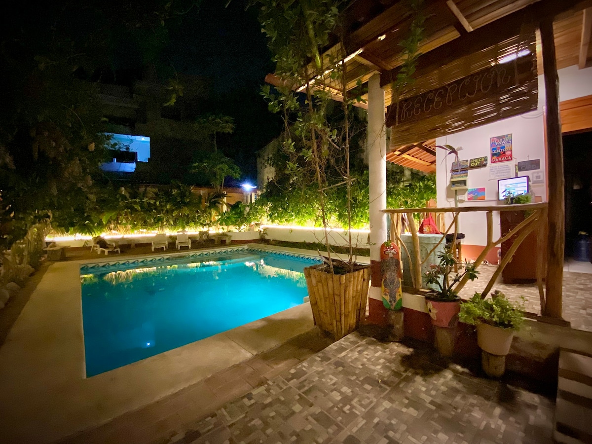 The outdoor area features a well-lit pool, surrounded by lush greenery and stone arrangements. A welcoming reception area is visible, with potted plants and decorative elements enhancing the space. Soft lighting creates a serene ambiance, inviting guests to enjoy the evening by the water.