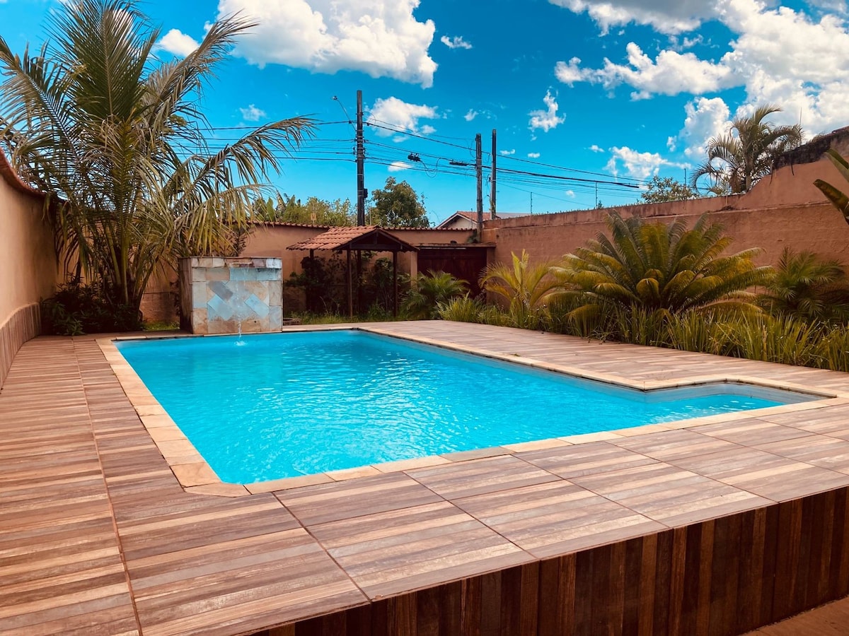 The image showcases a private swimming pool surrounded by wooden decking. Lush greenery flanks the pool area, with palm trees and shrubs adding a natural touch. A clear blue sky with scattered clouds is visible above, enhancing the outdoor ambiance.