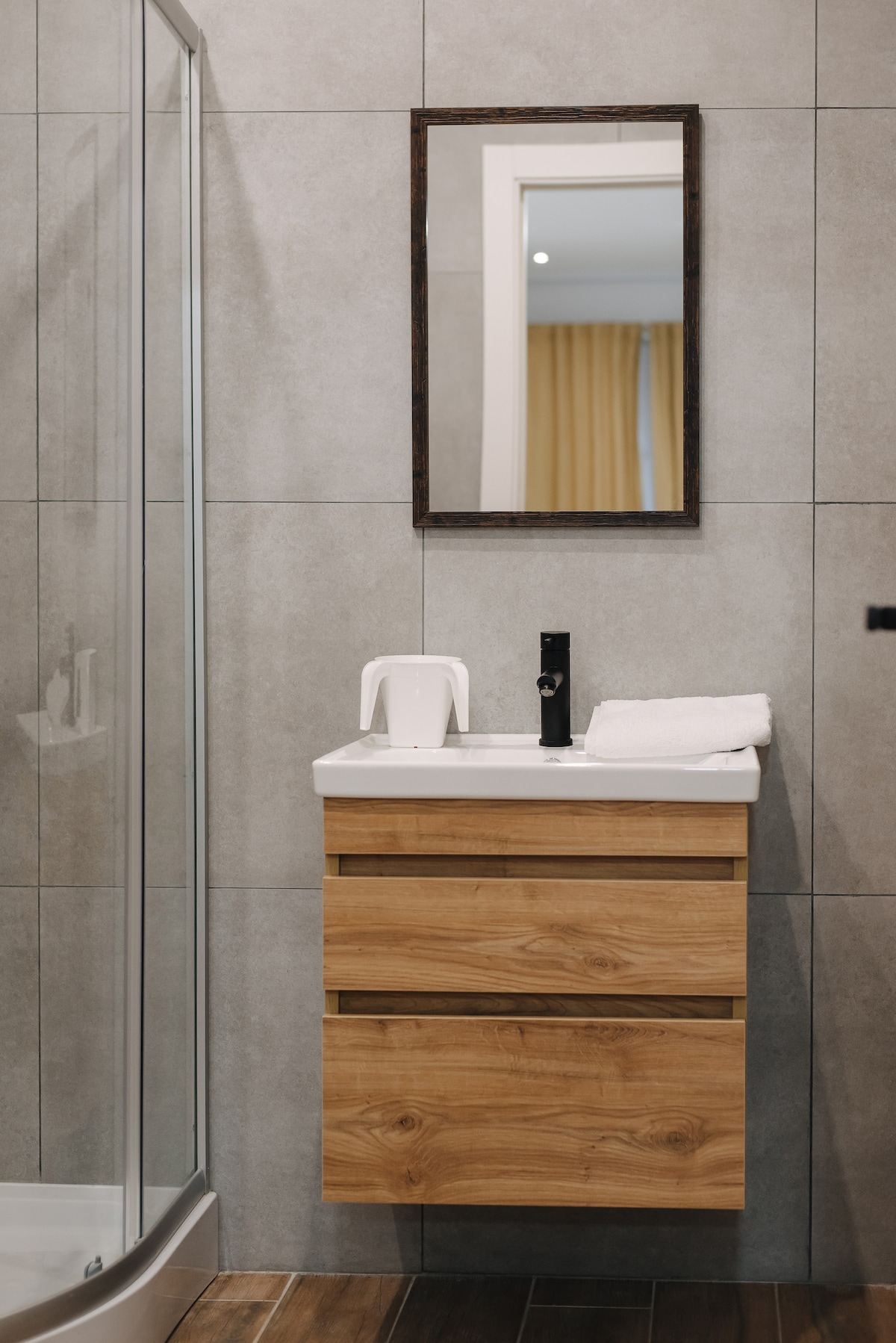A modern bathroom features a sleek wooden vanity with three drawers and a minimalist sink. A large mirror is mounted above the vanity, reflecting light from the interior. The shower area is enclosed with glass, and neutral tiles cover the walls.