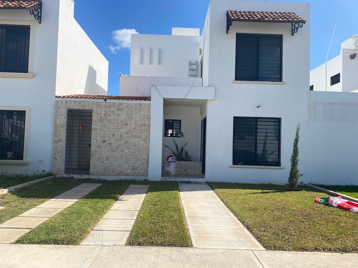 A two-story building features a modern facade, showcasing smooth white walls and decorative wrought iron elements. The front yard is neatly landscaped with green grass, bordered by paved walkways leading to the entrance. A festive decoration is visible in the garden area.