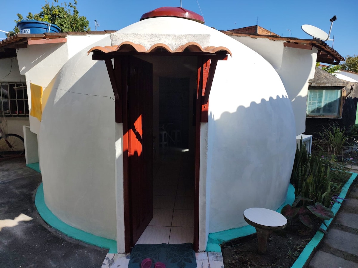 A unique dome-shaped structure is presented, featuring a smooth white exterior and a bright red entryway. The entrance showcases a simple wooden door with a curved awning. Surrounding greenery includes low plants and a small stone table positioned nearby, enhancing the inviting pathway.