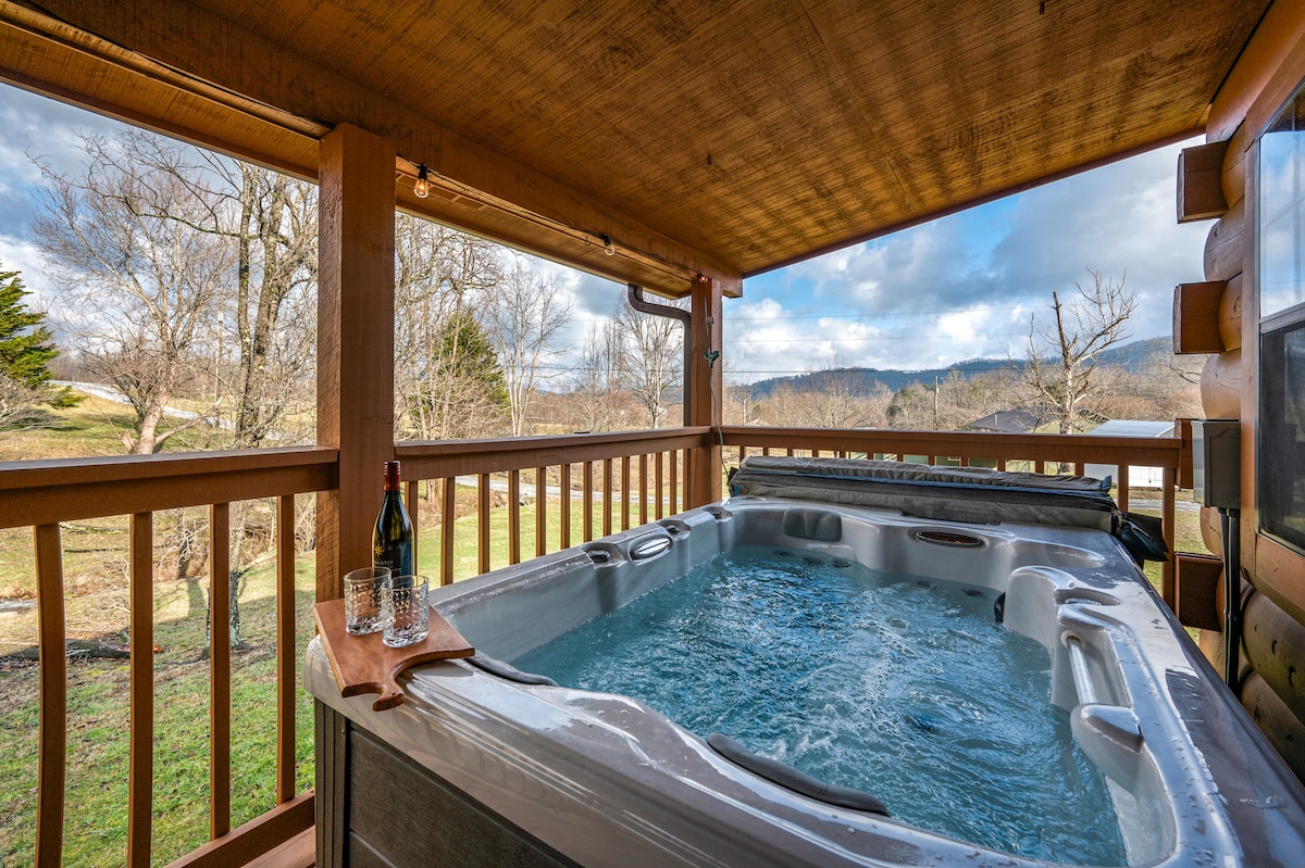 A private hot tub is positioned on a wooden deck, surrounded by a railing. Viewers can see a scenic landscape featuring grassy areas and trees in the background. Two glasses and a bottle of wine are placed on a adjacent wooden table.