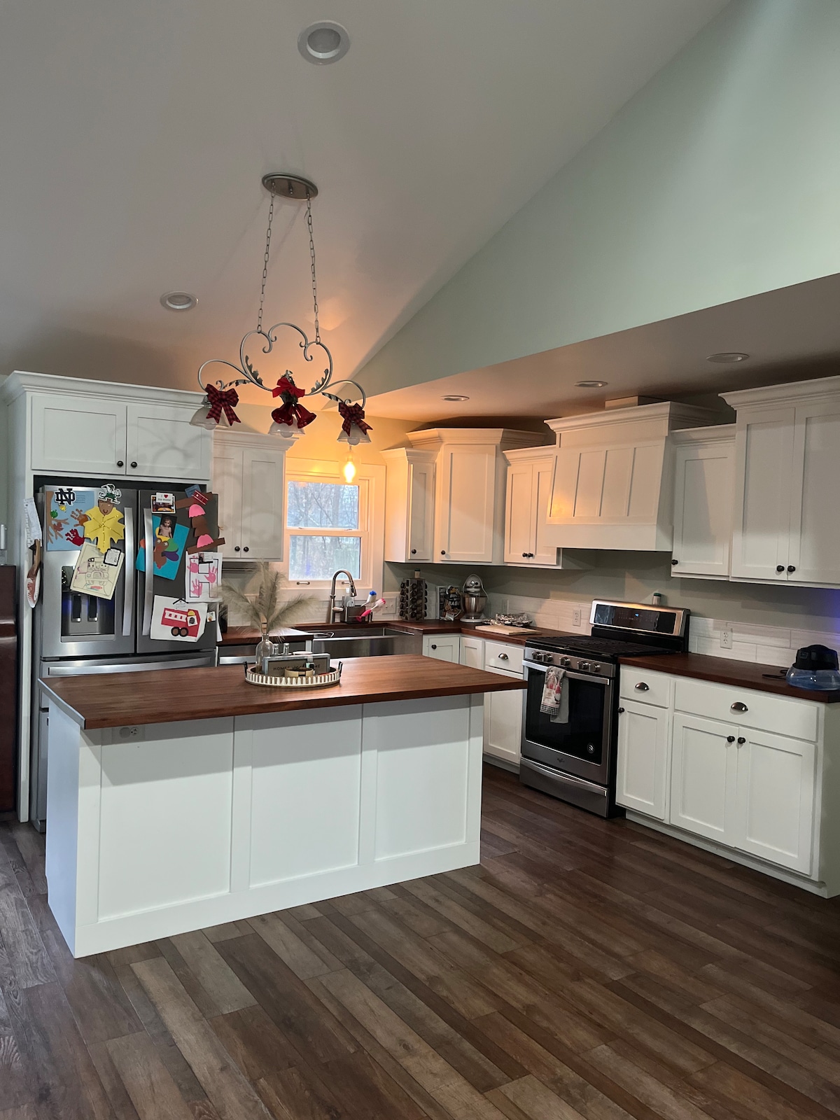 A bright kitchen features white cabinetry with ample storage space and a wooden island at the center. Stainless steel appliances are visible, including a refrigerator and an oven. A pendant light hangs above the island, illuminating the area, and windows allow natural light to enter.
