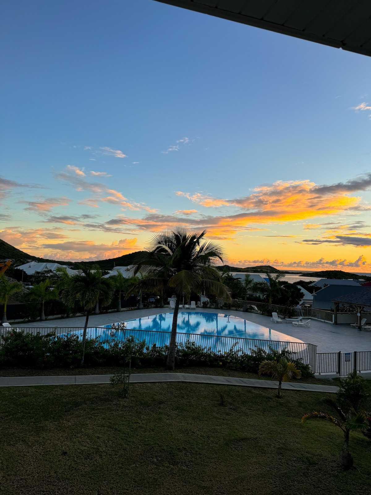 A serene evening view is displayed, featuring a large swimming pool surrounded by palm trees. The sky showcases vibrant hues as the sun sets, casting a warm glow over the tranquil scene. The area is bordered by residential buildings and lush greenery.