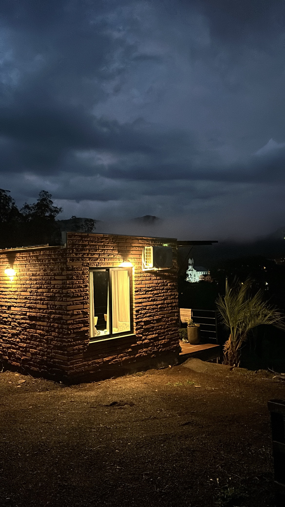 The stone cabin is illuminated by warm outdoor lights, surrounded by mist and dark clouds. A large window reveals light from within, contrasting with the evening sky. The landscape features hints of foliage and distant hills, adding to the serene atmosphere.