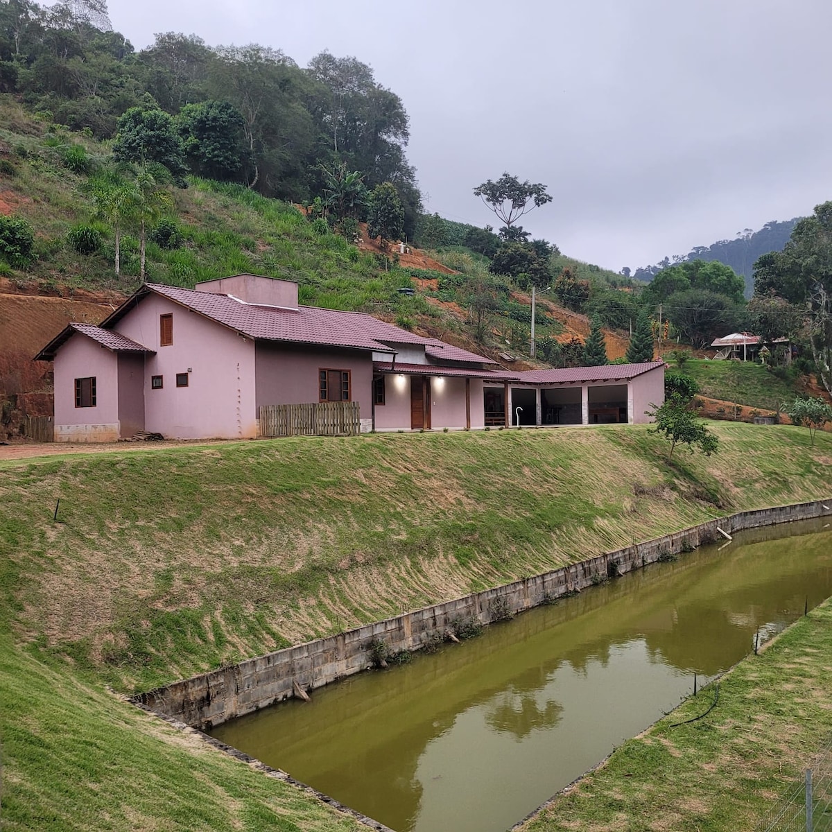 A pink house is set against a lush hillside, with greenery surrounding the property. A serene waterway runs in front, reflecting the natural landscape. The structure features a broad porch, complemented by a sloped roof and several windows that invite natural light.