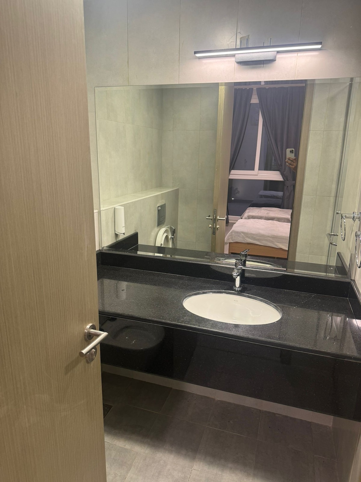 A modern bathroom featuring a sleek black countertop with an integrated sink. A large mirror reflects the space, while soft lighting provides adequate illumination. In the background, a glimpse of a bedroom is visible through an open doorway, showcasing a neatly made bed.