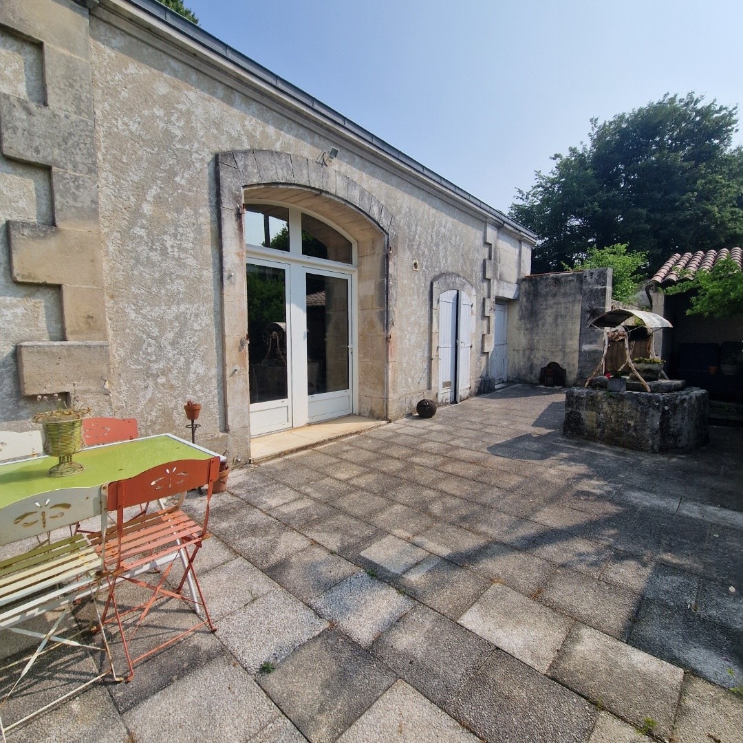 An outdoor patio area features a combination of rustic stone walls and tiled flooring. Two tables with colorful, mismatched chairs are positioned nearby. A stone fountain and lush greenery add to the relaxed ambiance, while sunlight bathes the space in warmth.