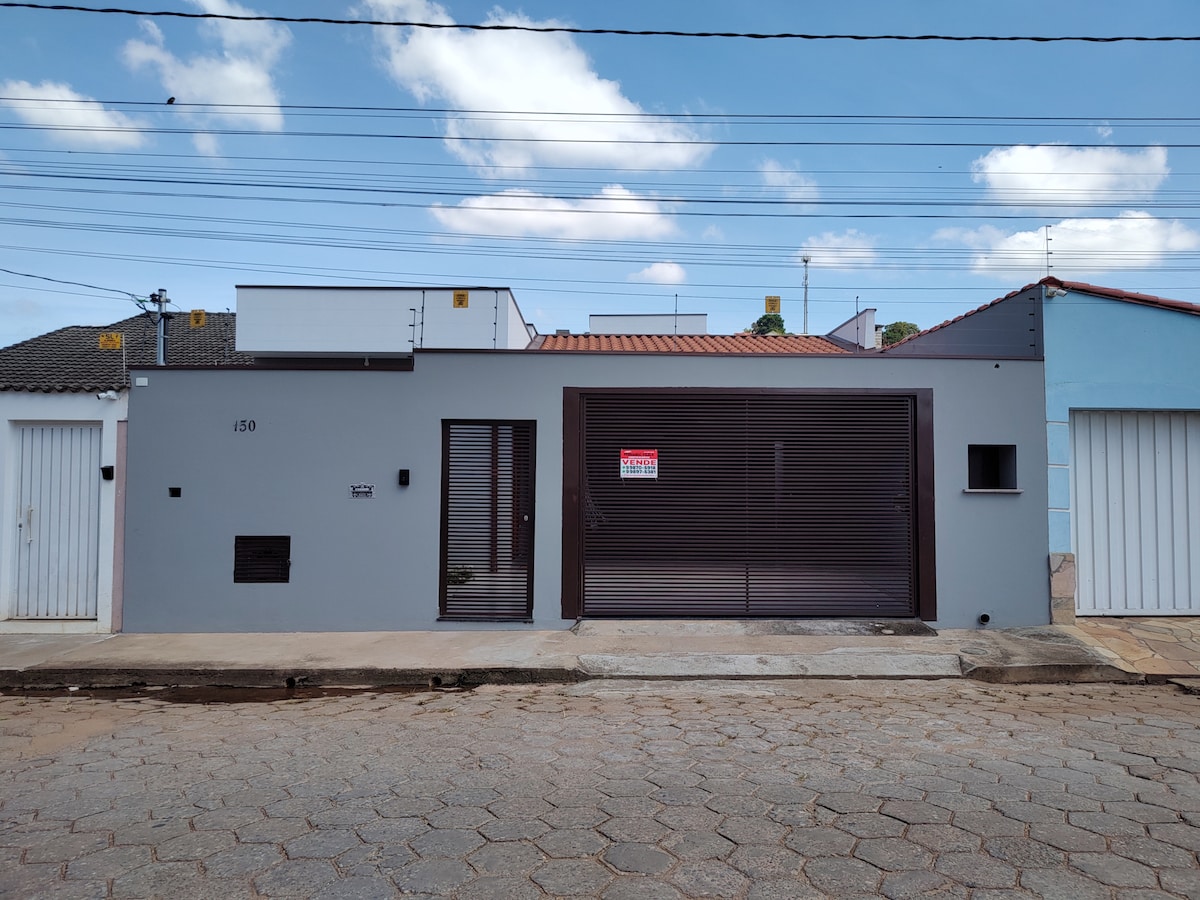 A single-story residence features a modern facade with a dark brown garage door and large windows. The exterior is painted in a light gray tone, contrasting with the earthy sidewalk. A clear blue sky is visible above, enhancing the structural lines of the property.