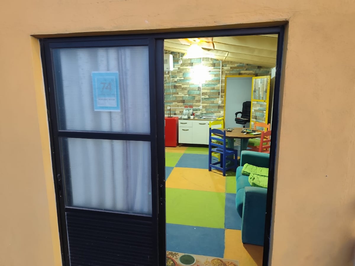 A view through the doorway reveals a vibrant living space with colorful patterned flooring. A cozy seating area is positioned alongside a dining table, complemented by a cheerful kitchen in the background. Natural light filters in, accentuating the lively atmosphere.